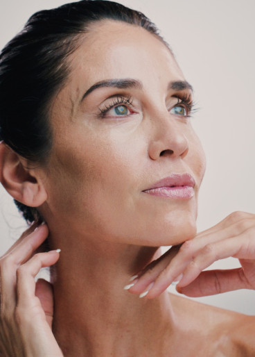 Eine schöne, zufriedene Frau mit dunklen Haaren, die nach einer Gesichtsbehandlung nachdenklich mit der Hand am Kinn hält und den Blick entspannt nach vorne richtet.